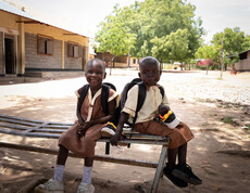 Zwei Grundschüler*innen mit Rucksäcken sitzen lachend auf Bank im Schulhof im Südsudan