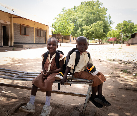 Zwei Grundschüler*innen mit Rucksäcken sitzen lachend auf Bank im Schulhof im Südsudan