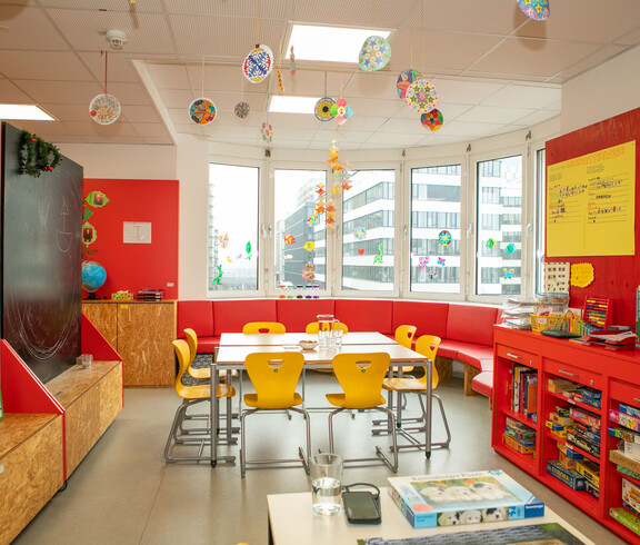 Bunter Lernraum mit Gruppentisch, Spielesammlung, Fensterdeko und roter Sitzecke im Caritas Lerncafé Wien.