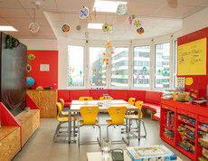 Bunter Lernraum mit Gruppentisch, Spielesammlung, Fensterdeko und roter Sitzecke im Caritas Lerncafé Wien.