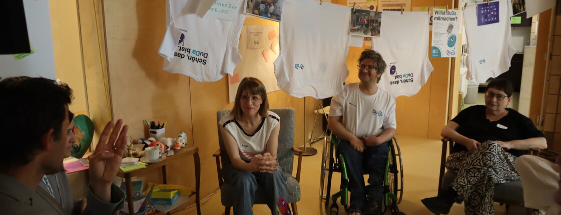 Mehrere Personen sitzen in einem Veranstaltungsraum im Kreis. Über der Gruppe hängen weiße T-Shirts und Fotos an einer Leine. Im Raum sind verschiedene Informationsmaterialien und Workshop-Utensilien sichtbar.
