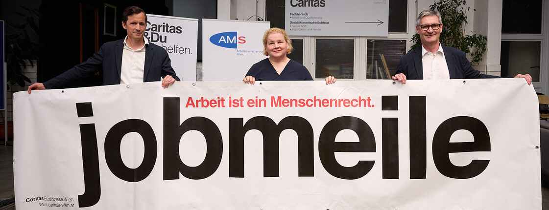 Drei Personen halten ein großes Banner mit der Aufschrift „jobmeile – Arbeit ist ein Menschenrecht“. Im Hintergrund sind Logos von Caritas und AMS sowie der Eingang eines Veranstaltungsraums zu sehen.