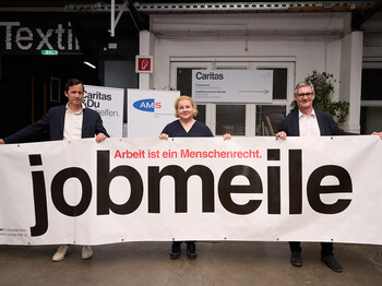 Drei Personen halten ein großes Banner mit der Aufschrift „jobmeile – Arbeit ist ein Menschenrecht“. Im Hintergrund sind Logos von Caritas und AMS sowie der Eingang eines Veranstaltungsraums zu sehen.