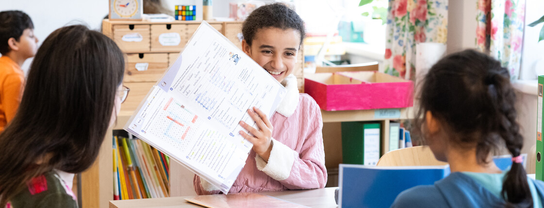 Eine Freiwillige und zwei Kinder sitzen mit Lernunterlagen an einem Tisch im Caritas Lerncafé