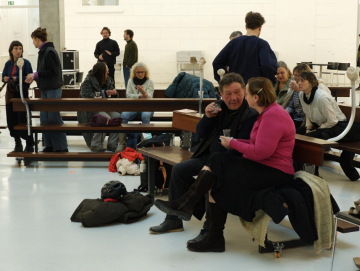 Menschen sitzen auf Bänken in einem hellen Saal, trinken Kaffee und unterhalten sich in kleinen Gruppen.
