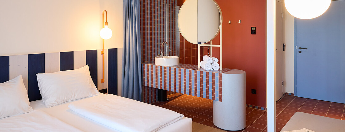 Innenansicht eines Hotelzimmers mit Doppelbett, integriertem offenem Waschbereich, rundem Spiegel, rot-blauen Fliesen, blauen Vorhängen und hellem Holzboden. Im Hintergrund befinden sich Eingangsbereich und Zimmertür.