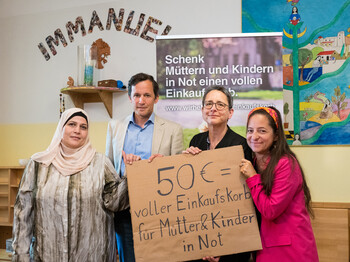 Personengruppe präsentiert ein Schild zur Spendenaktion „50 € = voller Einkaufskorb für Mütter & Kinder in Not“ in einem Gemeinschaftsraum.