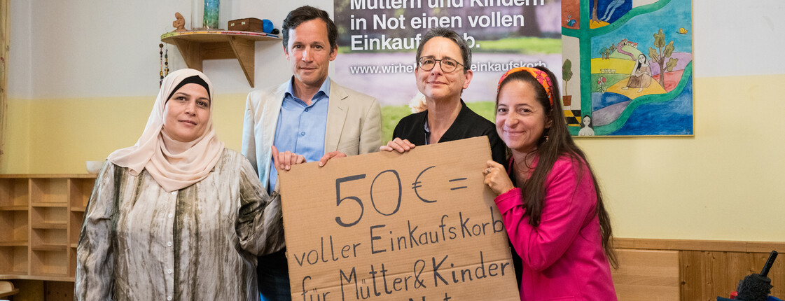 Personengruppe präsentiert ein Schild zur Spendenaktion „50 € = voller Einkaufskorb für Mütter & Kinder in Not“ in einem Gemeinschaftsraum.