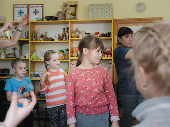Kinder stehen in einem Raum und tanzen. Im Hintergrund sind Regale mit Spielsachen und Deko-Gegenständen zu sehen.