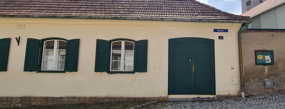Außenansicht älteres Gebäude, pastellgelbe Fassade mit grüner Holztür und weiße Holzfenster mit grünen Fensterläden aus Holz