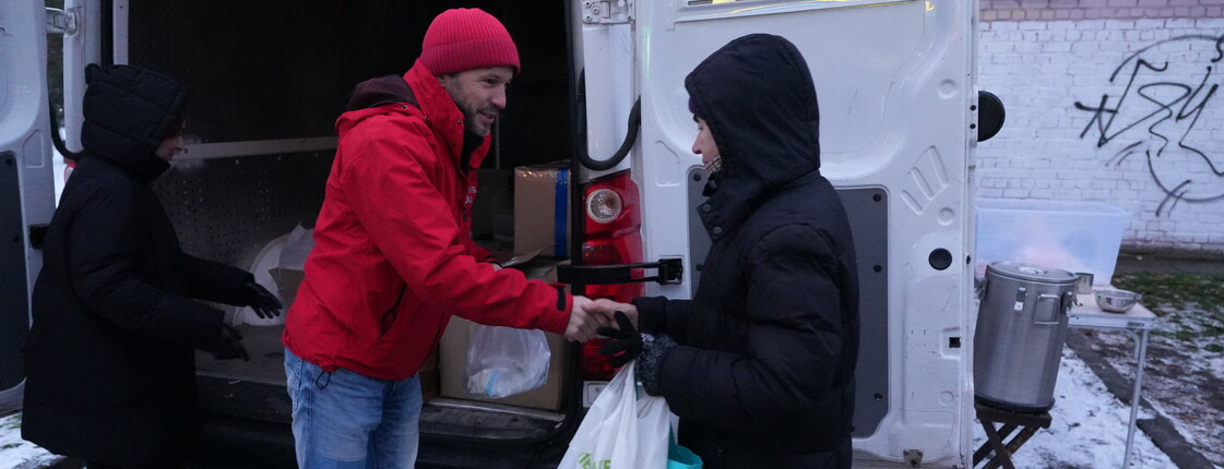 Personen verteilen Hilfsgüter aus einem geöffneten Lieferwagen. Eine Person im roten Mantel übergibt einer anderen eine Tüte mit Lebensmitteln. Im Hintergrund sind weitere Taschen, ein großer Topf auf einem Stativ und eine verschneite Umgebung zu sehen.