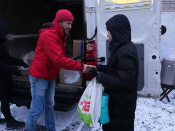 Personen verteilen Hilfsgüter aus einem geöffneten Lieferwagen. Eine Person im roten Mantel übergibt einer anderen eine Tüte mit Lebensmitteln. Im Hintergrund sind weitere Taschen, ein großer Topf auf einem Stativ und eine verschneite Umgebung zu sehen.