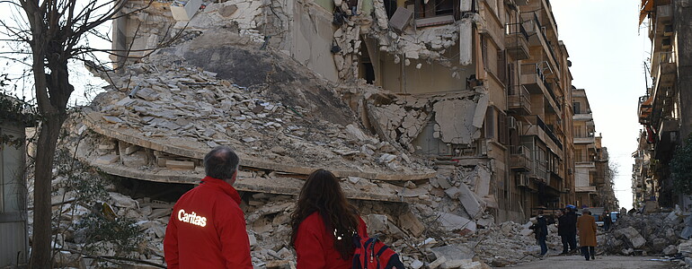zwei Mitarbeiter*innen der Caritas Österreich stehen vor eingestürzten Gebäuden in Aleppo