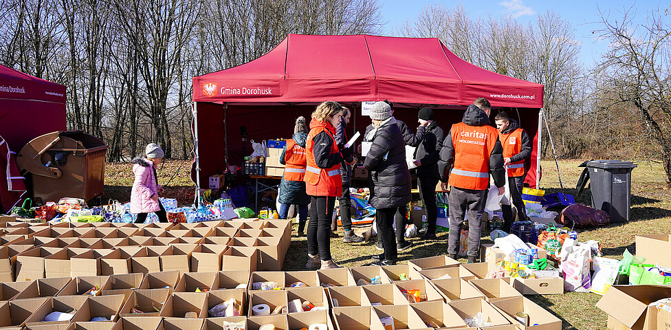 Man sieht ein Zelt, davor Menschen und Freiwillige der Caritas und viele Schachteln gefüllt mit Lebensmitteln und Lebensmitteln.